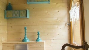 Une pièce en bois avec une porte en bois, une fenêtre ornée d’un rideau décoratif et deux étagères bleues fixées au mur.