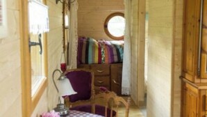 Un couloir étroit aux murs en bois, une fenêtre ronde, un lit avec une literie colorée, une chaise et une table avec une lampe.