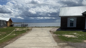 A concrete pathway leading to a wooden cabin with a metal roof, a small building with a brick facade, and a body of water in the background. - Lakefront Retreat At Randolphs Landing (Mannng)