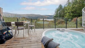 A hot tub on a wooden deck with a table and chairs, overlooking a scenic landscape with hills and a body of water.