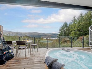 A hot tub on a wooden deck with a table and chairs, overlooking a scenic landscape with hills and a body of water. - Jack Strawbale House - UK7521 (Strontian, near Fort William)