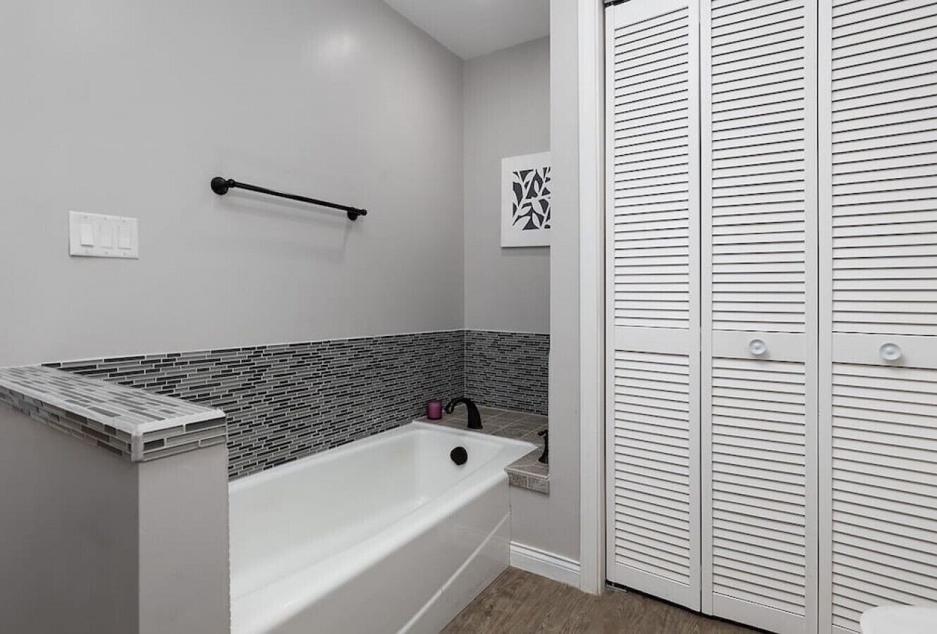 A modern bathroom with a white bathtub, a towel rack, a decorative tile backsplash, and a white door with louvered panels.