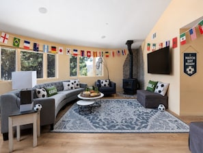Un salon avec un canapé gris aux lignes courbes, une table basse ronde sur laquelle sont disposés des en-cas, un téléviseur et une cheminée. Des drapeaux sont accrochés aux murs.