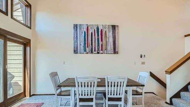 A dining area with a wooden table and white chairs, a colorful abstract painting on the wall, and a staircase to the right.