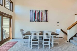 Un coin repas avec une table en bois et des chaises blanches, un tableau abstrait aux couleurs vives accroché au mur, et un escalier sur le côté droit.