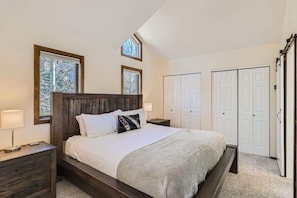 A bedroom with a wooden headboard, a bed with white bedding, two bedside tables with lamps, a large window, and a wardrobe.