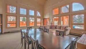 A spacious room with a large dining table surrounded by chairs, a sofa, and multiple large windows providing natural light.