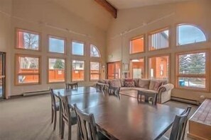 A spacious room with a large dining table surrounded by chairs, a sofa, and multiple large windows providing natural light.