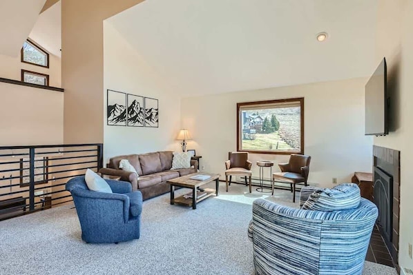 A spacious living room with a high ceiling, a fireplace, and a large window offering a view of the outdoors.