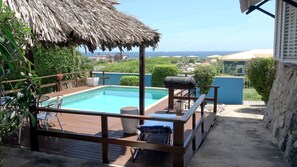 A pool area with a thatched roof, lounge chairs, and a barbecue grill.