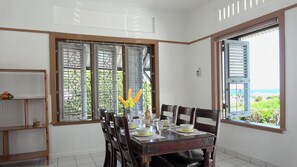 A dining area with a wooden table set for four, a vase with yellow flowers, and a view of greenery and a building through open windows.