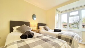 A bedroom with two beds, each with a checkered duvet, a nightstand with lamps, and a window with a view of the outside. - 2 bedroom accommodation in Winchester (Winchester)