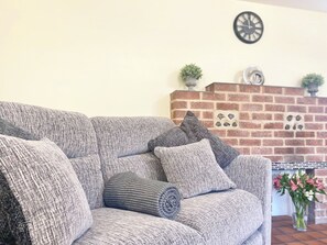 Un canapé gris recouvert de tissu, doté de coussins moelleux et d’un plaid texturé, placé devant une cheminée en briques agrémentée d’éléments décoratifs et surmontée d’une horloge murale.