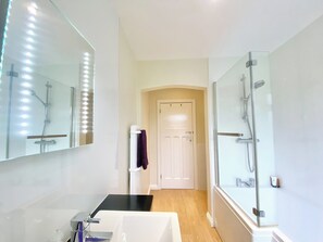 Un baño moderno con un espejo grande, un lavamanos blanco, cabina de ducha con paredes de vidrio y una puerta que da a otra habitación.