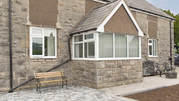 A stone building with a gabled roof, large windows, and a bench outside.