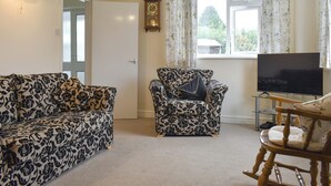 A living room with a sofa, armchair, and wooden rocking chair. There is a television on a stand and a clock on the wall.