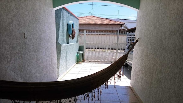 A hammock in a narrow corridor with tiled flooring, a white gate, and a building with a red roof in the background. - Vacation Home in Praia Grande - 3 min. Beach (Praia Grande)