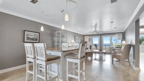 Una cocina amplia con taburetes blancos, una mesa de comedor, luces colgantes y vistas al exterior.