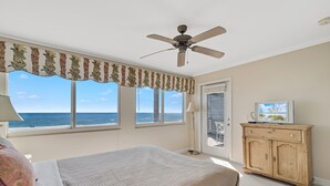 Un dormitorio con cama, ventilador de techo, aparador de madera y vista al océano a través de amplias ventanas.
