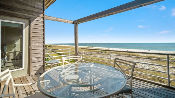 Terraza de madera con mesa y sillas de vidrio con vista a una playa.