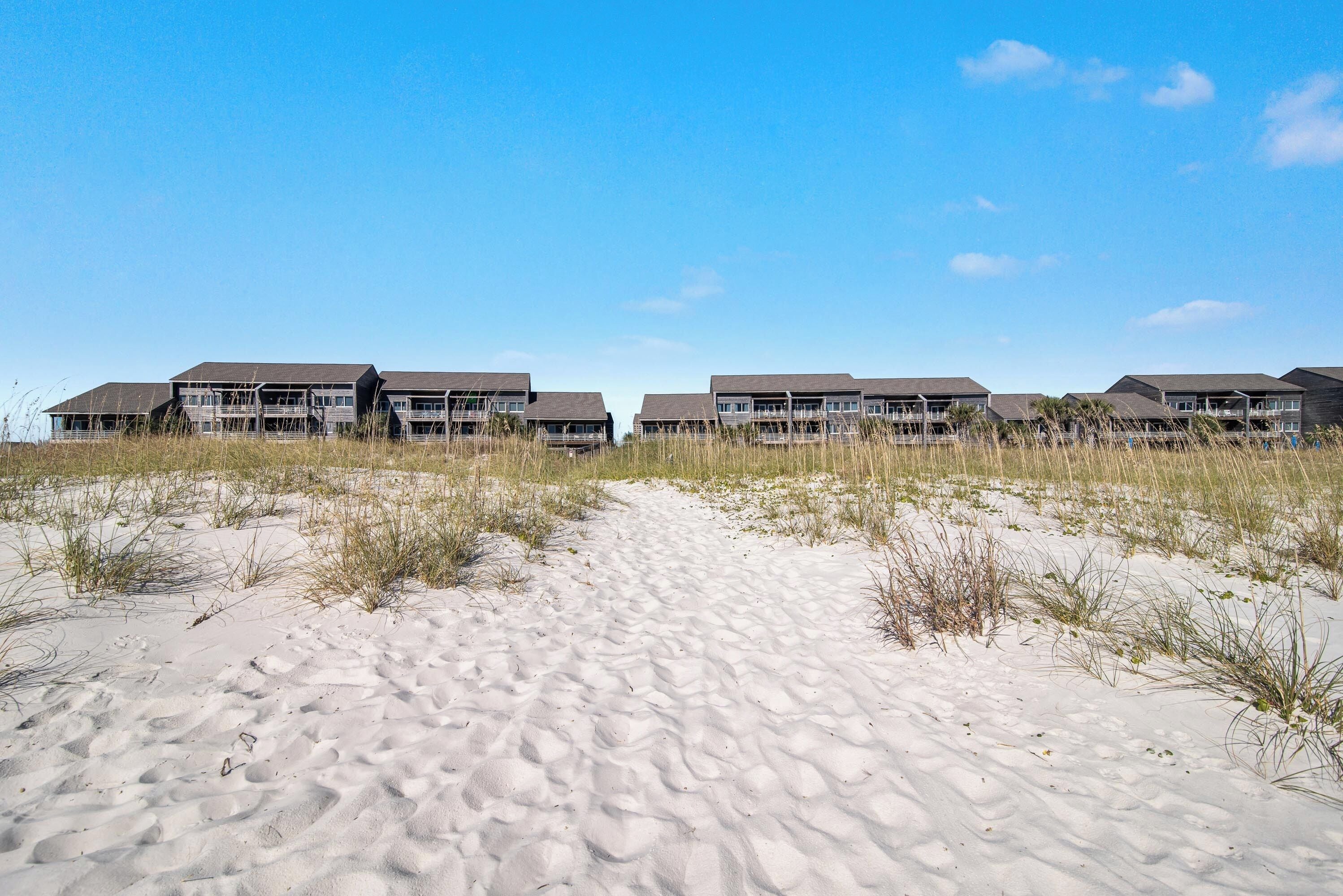 Un hôtel en bord de mer composé de plusieurs bâtiments, avec un chemin de sable menant à l’entrée et un ciel d’un bleu éclatant.