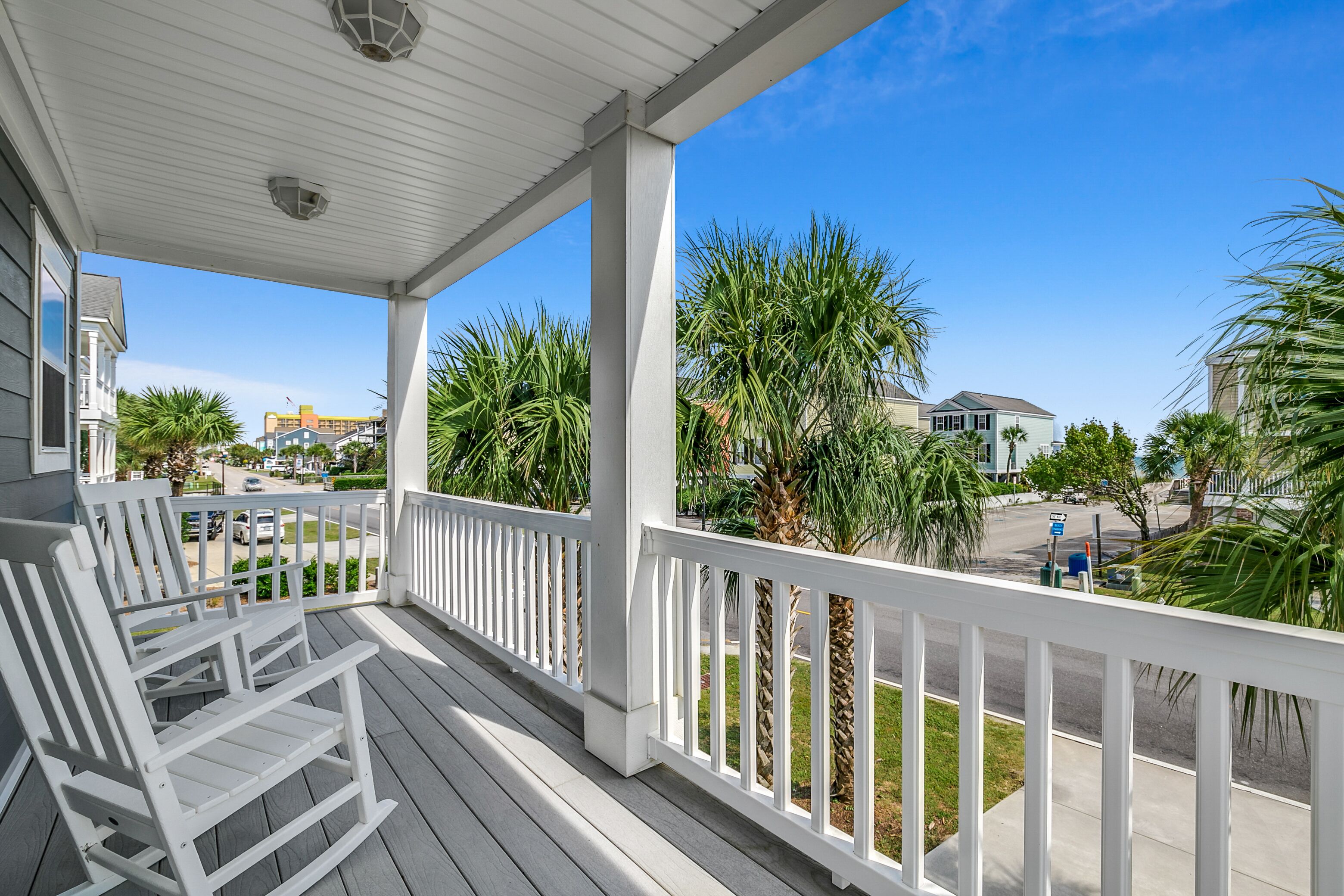A covered porch with white railings, a rocking chair, and a view of a residential street with palm trees and houses.