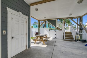 A covered patio with a wooden bench and a grill, a white door, and a staircase leading to a deck.