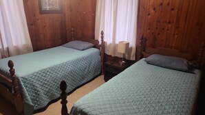 Ein Zimmer mit zwei hölzernen Betten, jeweils mit gesteppter Bettdecke und Kissen. Dazwischen befindet sich ein Nachttisch mit einer Lampe.