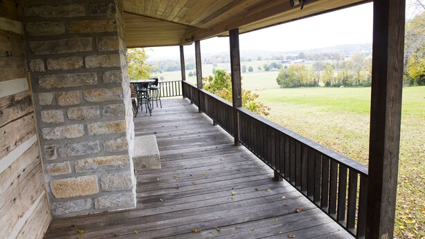A covered porch with stone walls, a wooden deck, and a view of a grassy field. - Historic Log Cabin on 200 acre farm, pasture and forest with panoramic views! (Lebanon)