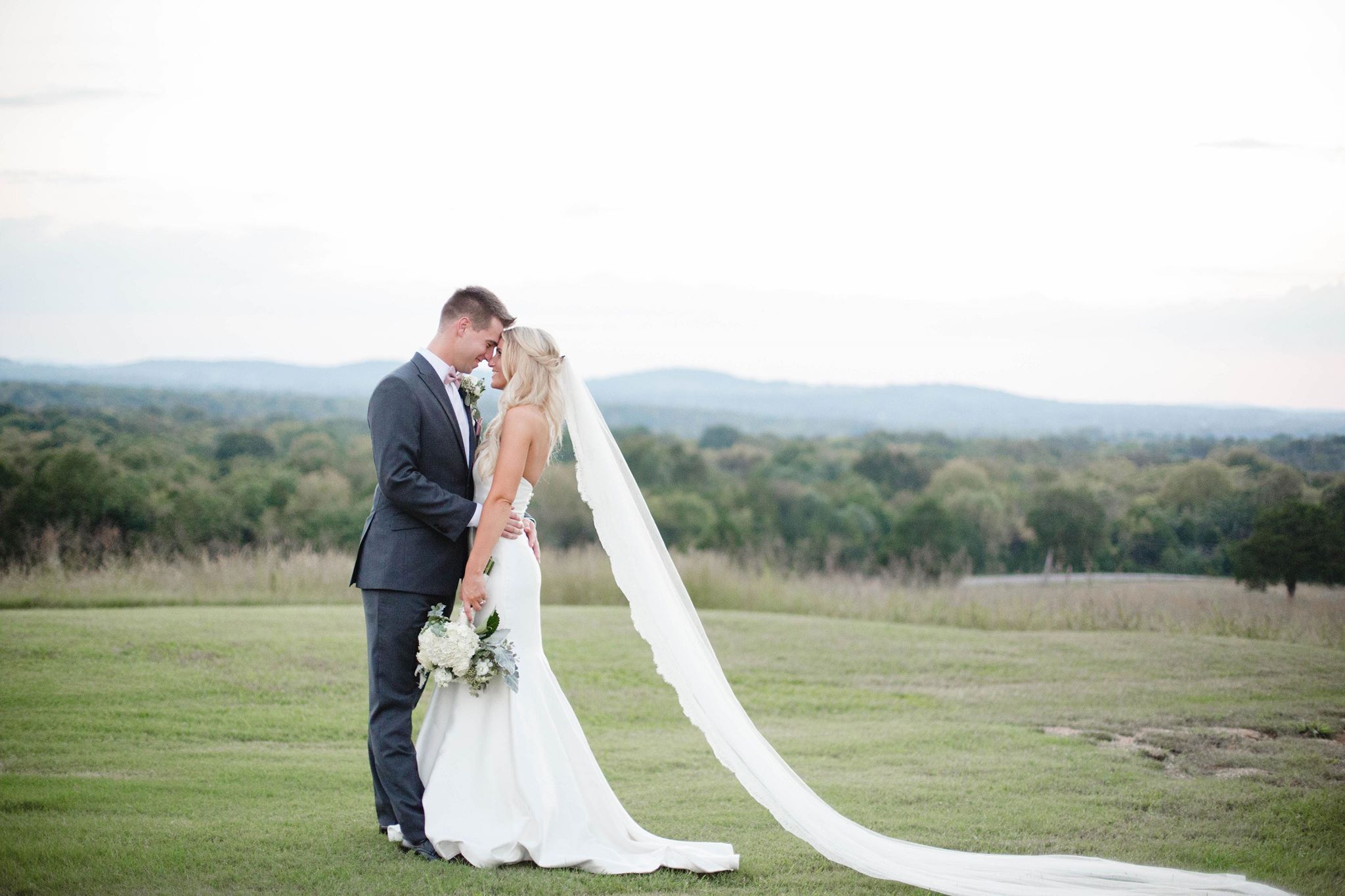 A couple standing on a grassy field with a mountainous backdrop.