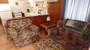 Un espacio habitable compacto con kitchenette, dos sillones estampados, una mesita y una lámpara.