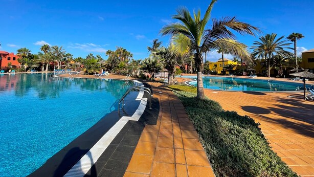 Un espace piscine aménagé dans un complexe hôtelier, avec une eau d’un bleu limpide, des palmiers et des chaises longues.