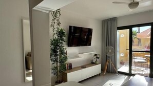 Ein modernes Wohnzimmer mit einem an der Wand befestigten Flachbildfernseher, einem weißen Sofa, einem kleinen Holznachttisch und einem Topf mit Pflanze.
