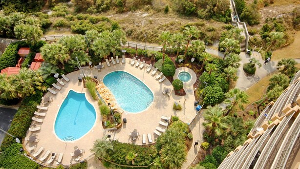 Vista aérea de un resort con dos piscinas, rodeado de palmeras y sillas de descanso.