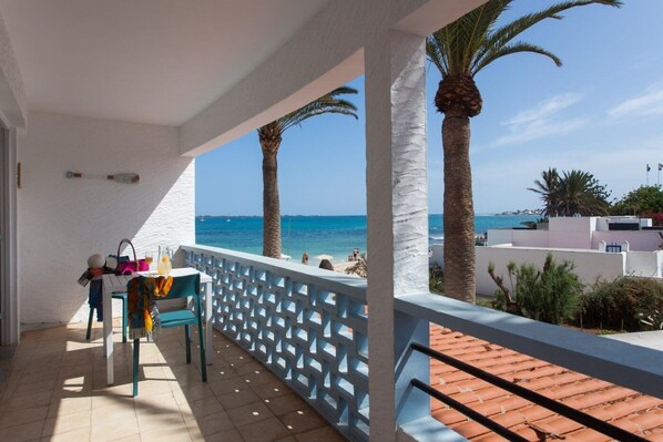 A balcony with a view of the sea, palm trees, and a tiled floor.