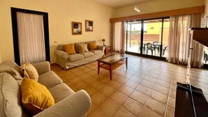 Una sala de estar con sofá, una mesa de centro y televisor. Hay dos cuadros enmarcados en la pared y un ventanal corredero que da a un espacio exterior con mesas y sillas.