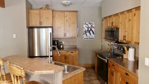 A kitchen with wooden cabinets, stainless steel appliances, and a small island with bar stools. - Ground Level, 2 Bedroom Suite - Close to Village and The Pond!  (Silver Star Mountain)