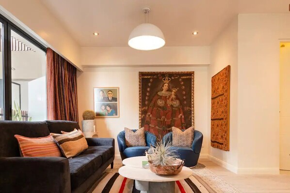 A modern living room with a dark sofa, blue armchair, and a round coffee table. A large tapestry hangs on the wall, and there are framed pictures on the wall.