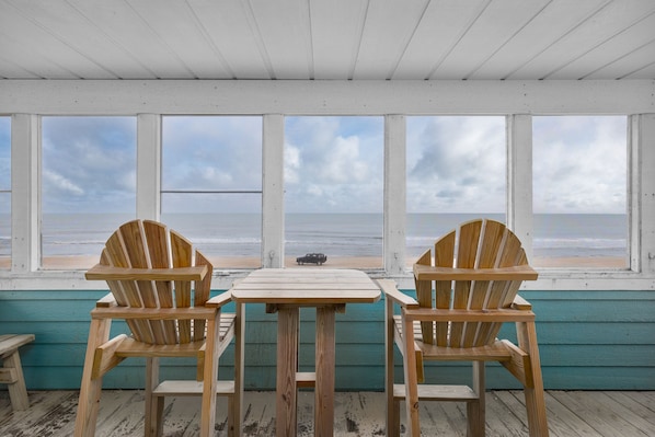A wooden deck with two Adirondack chairs and a small table, overlooking a beach with a cloudy sky.