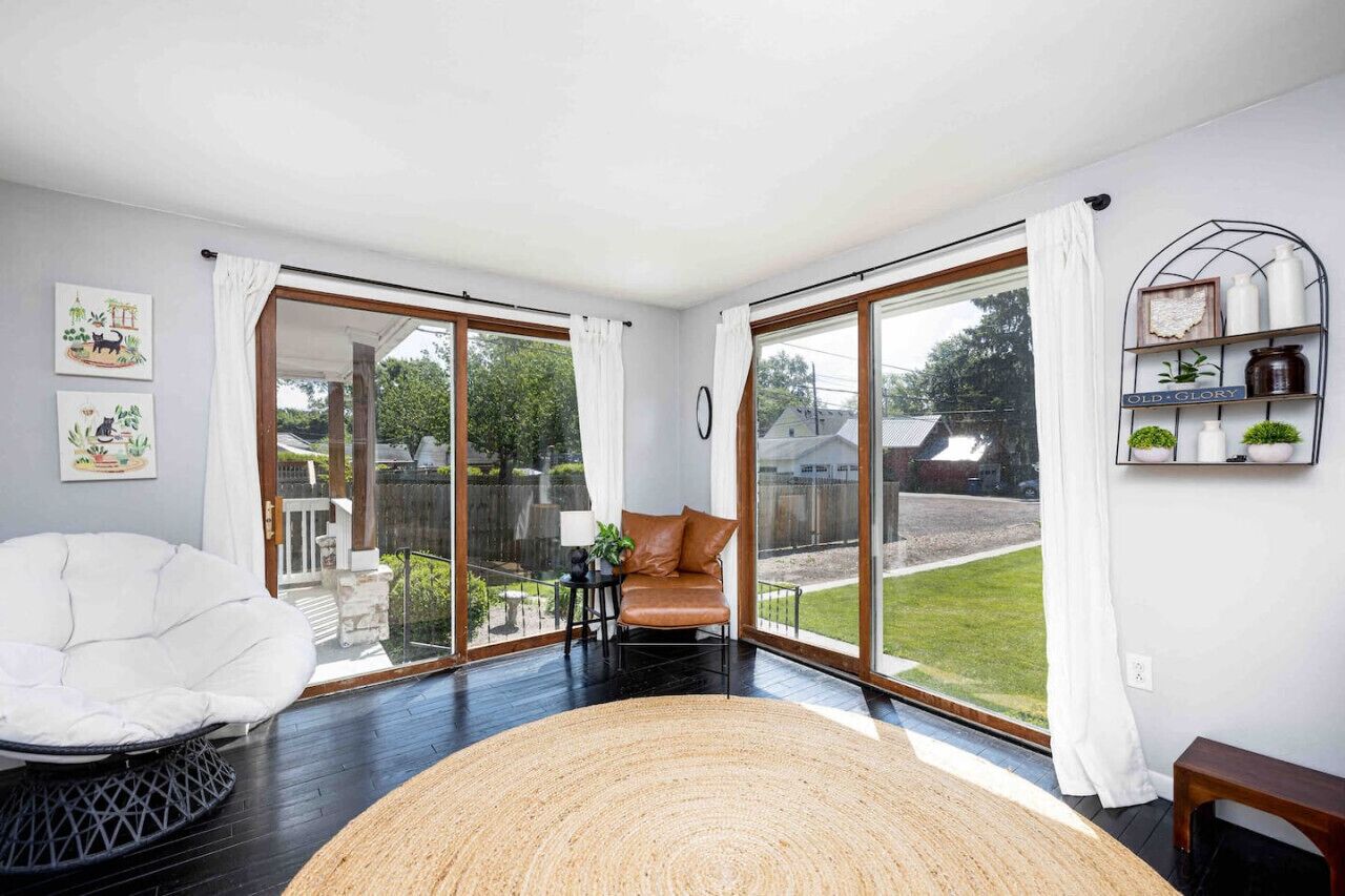 Un salon moderne doté d’une grande porte-fenêtre coulissante donnant sur un jardin, d’un fauteuil blanc, d’un tapis rond en fibres naturelles et d’une étagère ornée d’objets décoratifs.