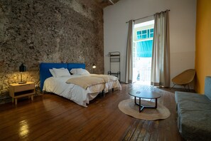 Un dormitorio con pared de piedra, una cama con cabecera azul, una mesita de noche, una mesa redonda y una silla.