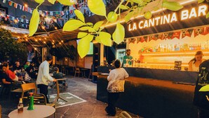 Zona de bar al aire libre con mesas y sillas, una barra con un cantinero, y un letrero que dice 'LA CANTINA BAR'.