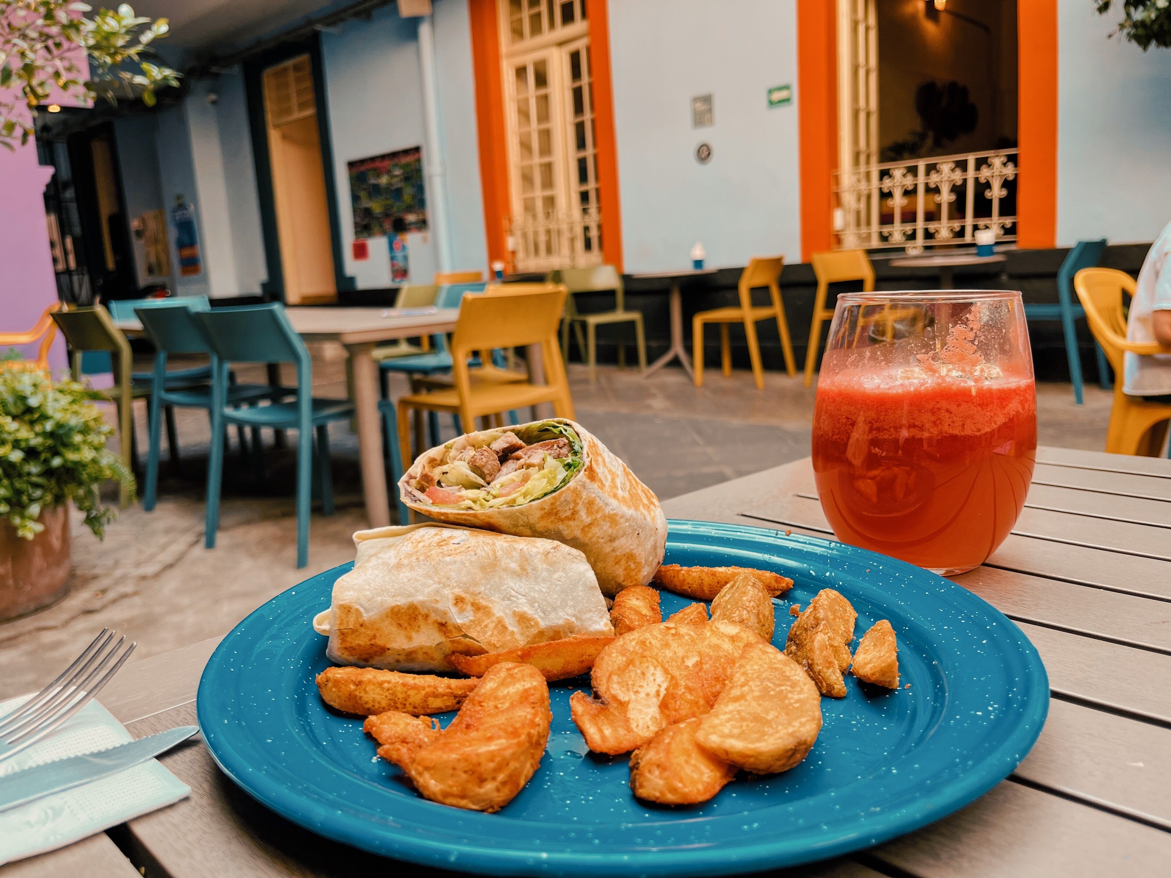 Un plato con un wrap y alimentos fritos, un vaso de bebida roja, y mesas y sillas al aire libre.