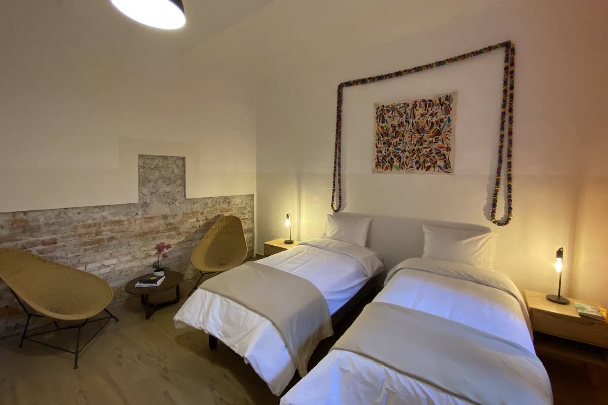 Hotellihuone, jossa on kaksi sänkyä, punottu tuoli, pieni pöytä, seinälle kiinnitetty taideteos ja yksi seinä, joka on koristeltu tiilillä.