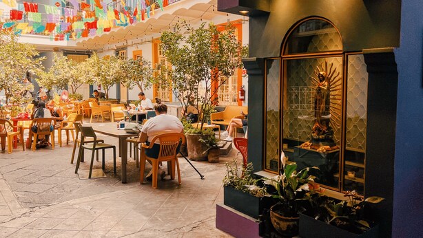 Comedor interior con mesas y sillas, plantas en macetas y un ventanal decorativo con una estatua.