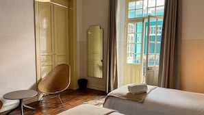 Habitación de hotel con dos camas, una mesa redonda, una silla de mimbre, un espejo y una ventana con cortinas.