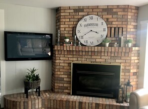 Una sala de estar con chimenea de ladrillo, un reloj montado en la pared y un televisor de pantalla plana.
