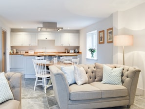 Una cocina moderna con armarios blancos, una zona de comedor con mesa de madera y sillas blancas, y un sofá beige con cojines estampados.
