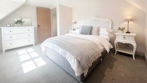 Quarto com teto inclinado, cama com cabeceira branca, mesa de cabeceira com abajur e cômoda.