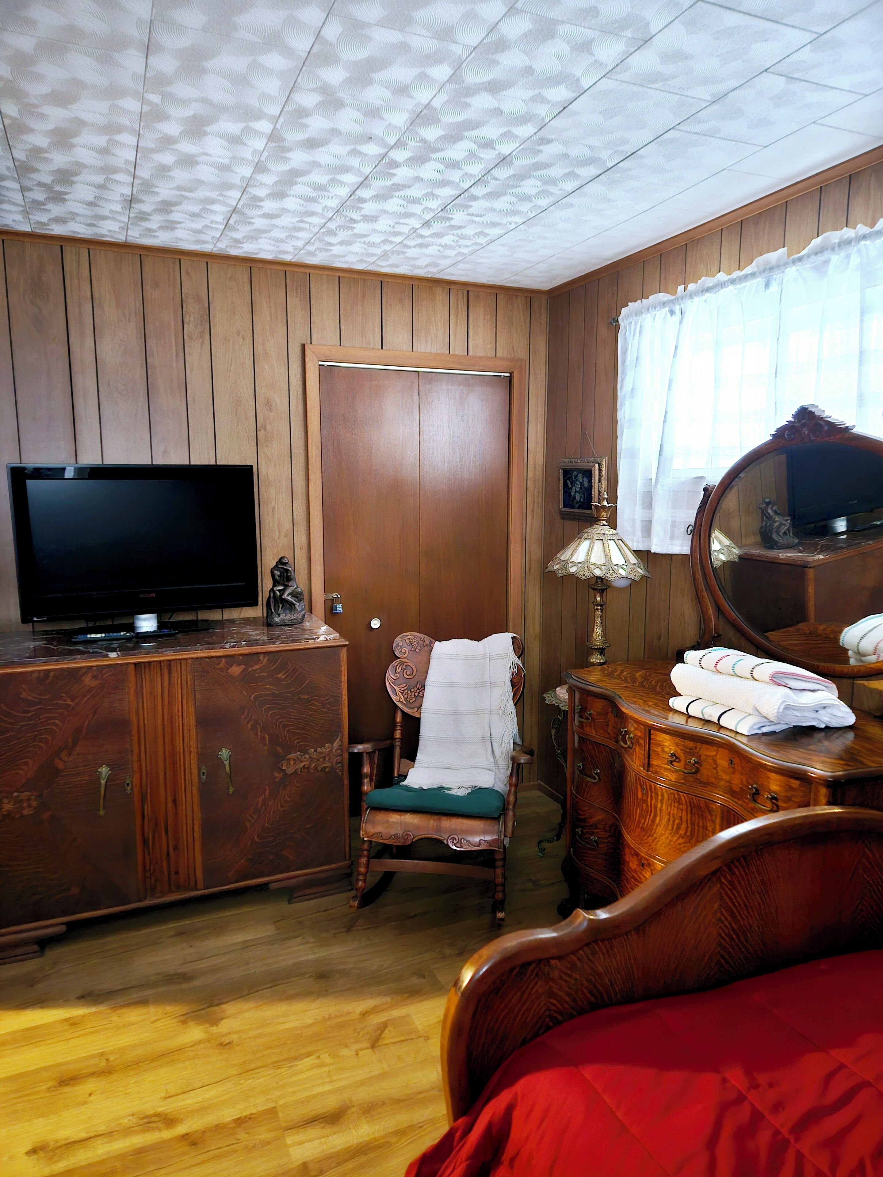 A room with a wooden wardrobe, a television, a chair with a blanket, a dresser with a mirror, and a bed.